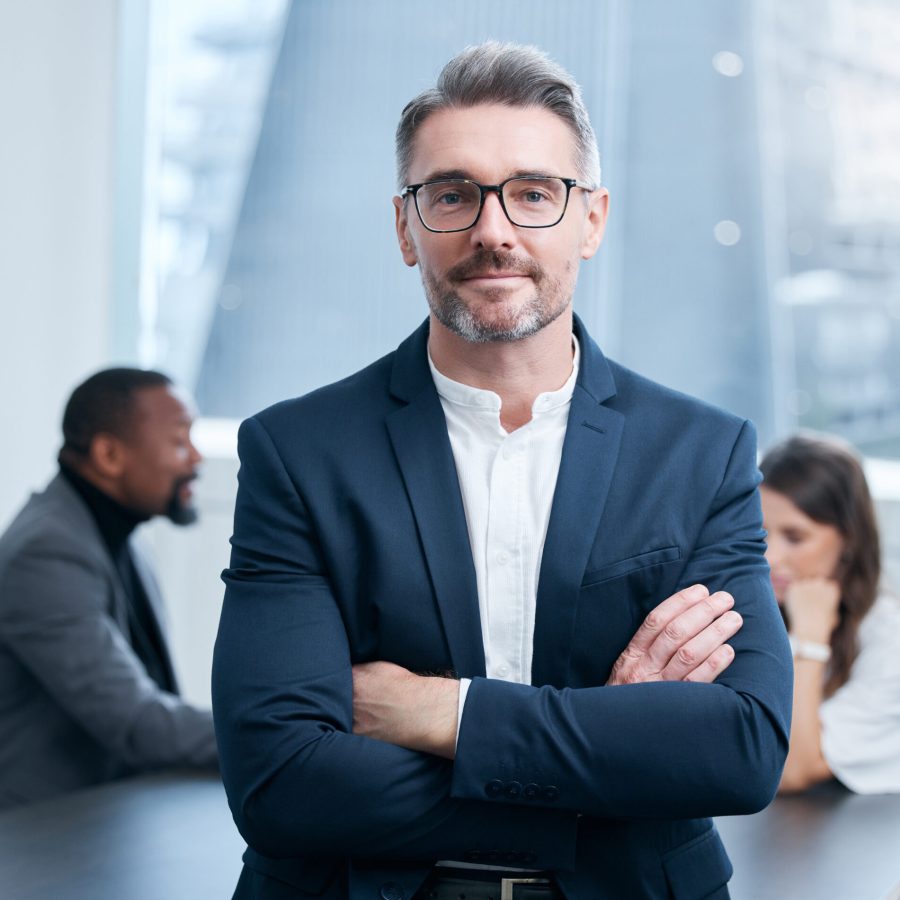 Portrait of a confident mature businessman standing in an office with his colleagues in the background.